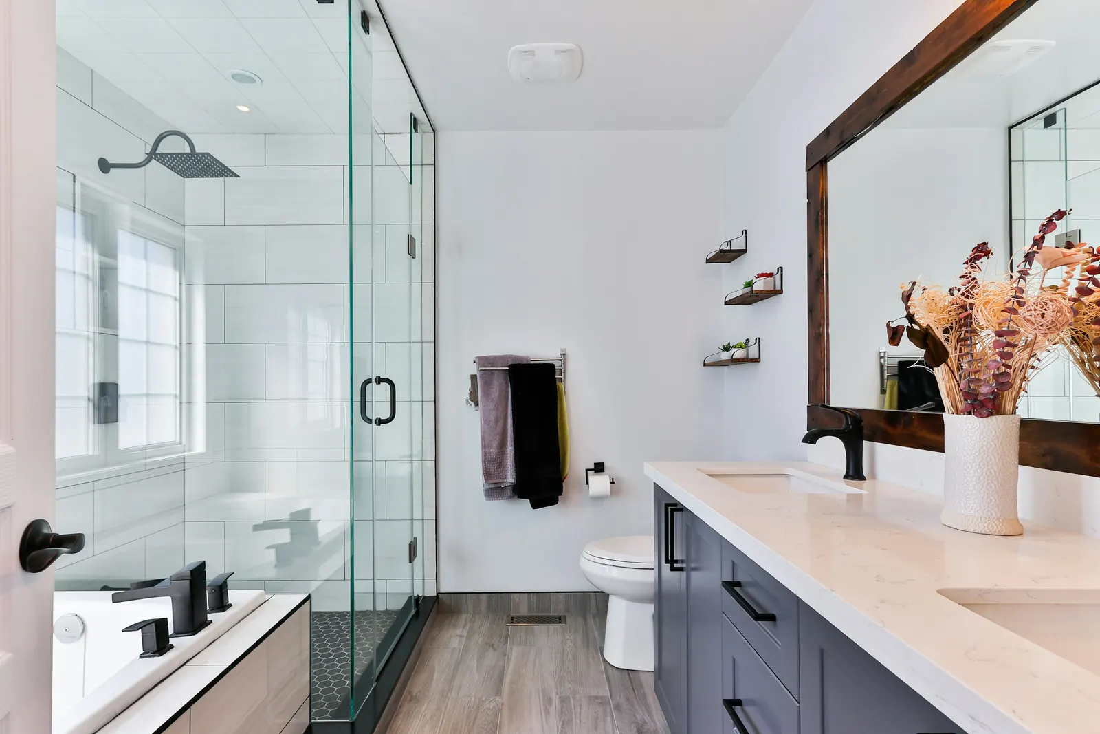A bathroom with a glass shower and a double vanity under a wood-framed mirror